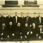 16 men in suits stand pose for a picture outside of a courthouse in a sepia photo.