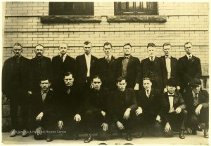 16 men in suits stand pose for a picture outside of a courthouse in a sepia photo.