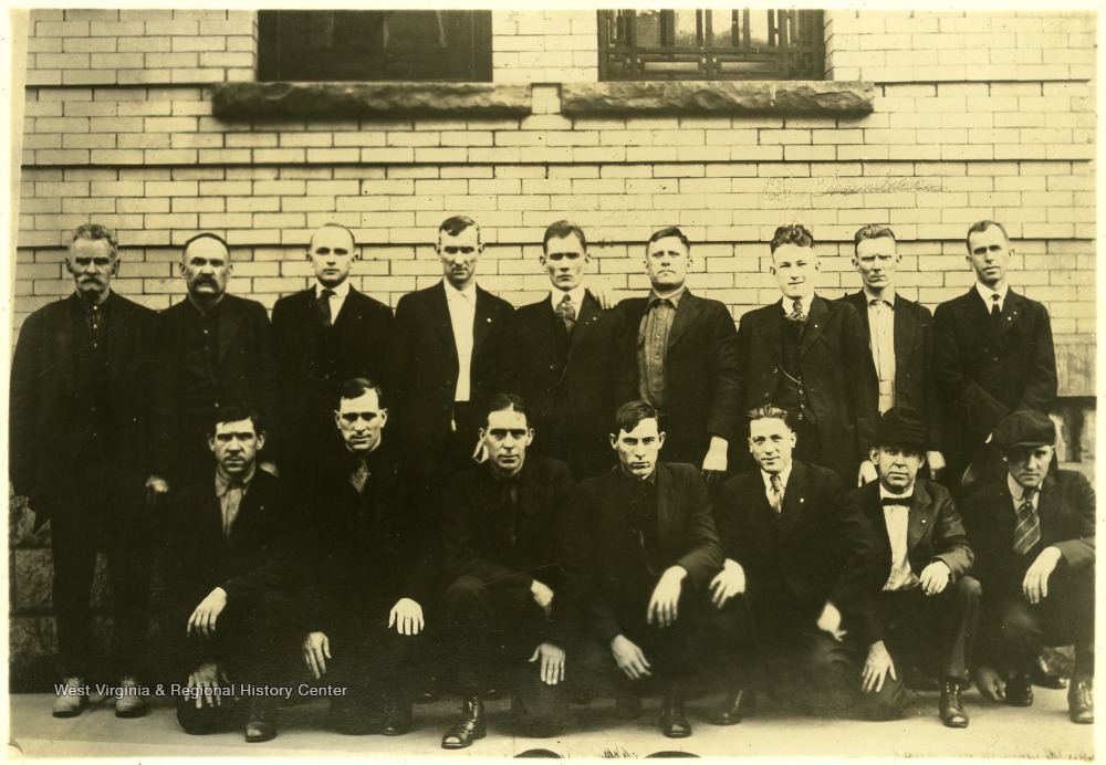 16 men in suits stand pose for a picture outside of a courthouse in a sepia photo.