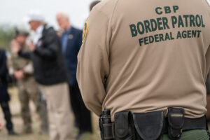 Customs and Border Patrol (CBP) agent's back. In the background there is several men talking out of focus. 