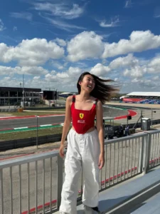 A joyful woman in white pants and a red top stands by a racetrack fence, her hair blowing in the wind under a sunny sky with clouds.