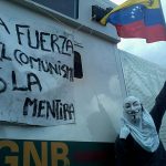 A protester wearing an Anonymous mask and holding a Venezuelan flag. The sign says "The strength of communism is the deceit". March 16, 2014